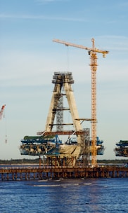 A partially constructed bridge visible over a body of water, featuring a prominent A-shaped structure being supported by cranes. The construction is ongoing with scaffolding and machinery, surrounded by a clear sky.