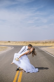 A couple is captured in an affectionate embrace on an open road, with the bride being dipped slightly backward by the groom. The bride is wearing a flowing white wedding dress and holding a bouquet of flowers. The groom is dressed in a white shirt with suspenders. The surroundings include an expansive, deserted highway with a clear blue sky above and dry, arid landscape in the background.