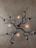 An artistic light fixture is mounted on a wall. The design resembles twisted, bare tree branches or vines with small, ornamental elements. Several light bulbs are integrated into the branches, casting a warm glow against the neutral-colored wall.