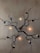 An artistic light fixture is mounted on a wall. The design resembles twisted, bare tree branches or vines with small, ornamental elements. Several light bulbs are integrated into the branches, casting a warm glow against the neutral-colored wall.