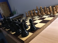 A wooden chessboard is set up for a game with black and white pieces positioned in their starting places. The board is placed on a wooden table, and there is soft lighting in the room. In the background, papers are loosely stacked on a table.