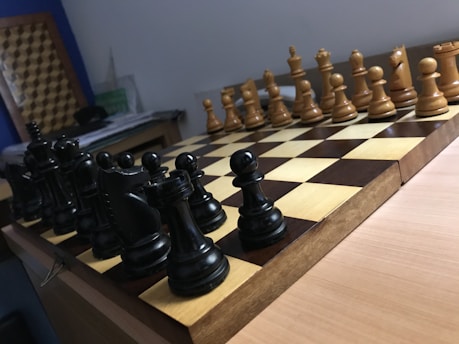 A wooden chessboard is set up for a game with black and white pieces positioned in their starting places. The board is placed on a wooden table, and there is soft lighting in the room. In the background, papers are loosely stacked on a table.