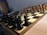 A wooden chessboard is set up for a game with black and white pieces positioned in their starting places. The board is placed on a wooden table, and there is soft lighting in the room. In the background, papers are loosely stacked on a table.