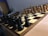 A wooden chessboard is set up for a game with black and white pieces positioned in their starting places. The board is placed on a wooden table, and there is soft lighting in the room. In the background, papers are loosely stacked on a table.