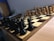 A wooden chessboard is set up for a game with black and white pieces positioned in their starting places. The board is placed on a wooden table, and there is soft lighting in the room. In the background, papers are loosely stacked on a table.