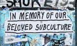 A large poster with bold black lettering reads 'In Memory of Our Beloved Subculture' pasted on a wall covered in graffiti and various stickers. The background shows a mix of blue and teal tones with visible street art and advertisements.