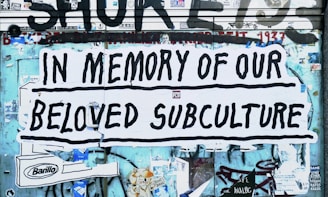 A large poster with bold black lettering reads 'In Memory of Our Beloved Subculture' pasted on a wall covered in graffiti and various stickers. The background shows a mix of blue and teal tones with visible street art and advertisements.