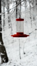 A red bird feeder suspended from a chain stands out against a snowy woodland background with bare, snow-covered tree branches.