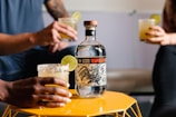 A bottle of Espolon tequila is placed on a yellow geometric table, flanked by two people holding glasses with salted rims and lime garnishes. The focus is on the bottle, which features intricate black and white artwork on the label.