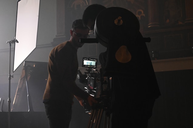 A dimly lit setting featuring two individuals working with film equipment, including a large camera on a tripod and lighting equipment. The environment suggests a focus on film production or cinematography.
