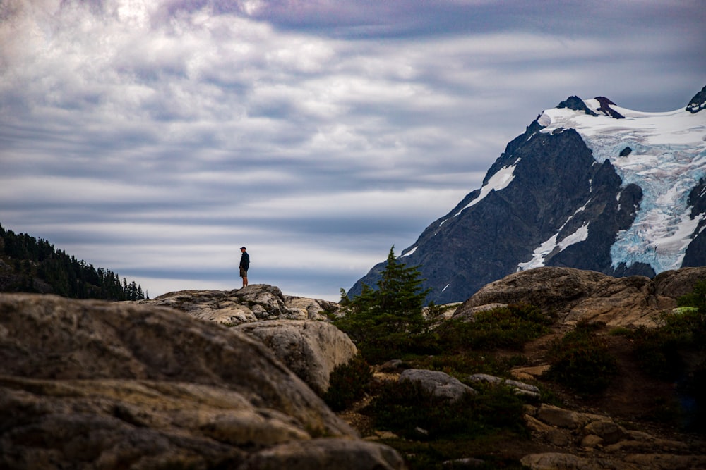 Person, die tagsüber unter bewölktem Himmel auf einem Felsen in der Nähe des Berges steht