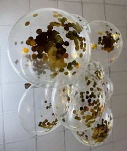 Transparent balloons filled with gold confetti cluster together against a tiled ceiling. The light reflects off the confetti, creating a shimmering effect.