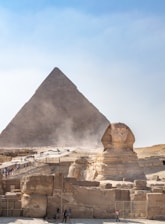 A large pyramid and the Great Sphinx against a backdrop of a clear sky. Several tourists are present, walking along designated paths.