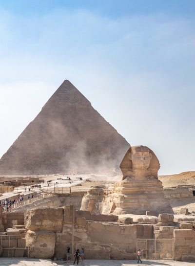 A large pyramid and the Great Sphinx against a backdrop of a clear sky. Several tourists are present, walking along designated paths.