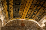 An ornate ceiling filled with intricate Ancient Egyptian hieroglyphs and symbolic figures, predominantly in shades of yellow, black, and blue. The artwork is detailed, suggesting scenes and deities, with the presence of figures like serpents and human-like shapes. The ceiling illustrates a historical and mythological narrative.