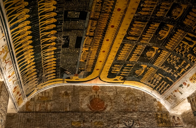 An ornate ceiling filled with intricate Ancient Egyptian hieroglyphs and symbolic figures, predominantly in shades of yellow, black, and blue. The artwork is detailed, suggesting scenes and deities, with the presence of figures like serpents and human-like shapes. The ceiling illustrates a historical and mythological narrative.