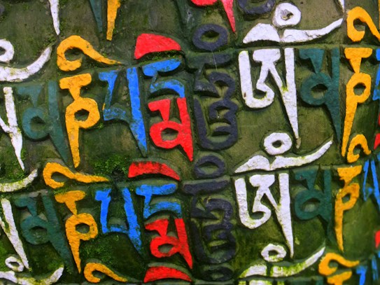 A colorful and intricate stone surface featuring carved Tibetan or Sanskrit script painted in vivid shades of white, yellow, red, blue, and green. The script is densely arranged, creating a textured and vibrant pattern.