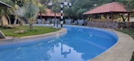 A serene swimming pool with clear blue water curves around a landscaped area with grass and palm trees. Several thatched huts with red-tiled roofs and outdoor seating arrangements are visible in the background, surrounded by lush greenery. A stone pathway runs beside the pool, and a lamp post stands near the edge.