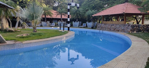 A serene swimming pool with clear blue water curves around a landscaped area with grass and palm trees. Several thatched huts with red-tiled roofs and outdoor seating arrangements are visible in the background, surrounded by lush greenery. A stone pathway runs beside the pool, and a lamp post stands near the edge.