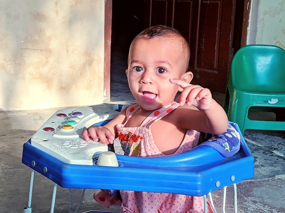 A baby in a pink outfit is sitting in a blue baby walker with a colorful control panel. The baby is pointing with one hand while the mouth is slightly open, creating an expression of curiosity or excitement. The room has a rough textured cream-colored wall, a wooden door in the background, and a green plastic chair to the side.