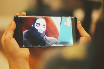A smartphone is being held by a person, displaying an image of a character wearing a black mask with a white skull design. The background features bright colors, including red and blue.