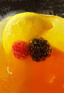 A close-up view of a chilled drink containing a slice of lemon, a red berry, and a black berry submerged in liquid. Bubbles cling to the surface of the fruits, indicating carbonation.