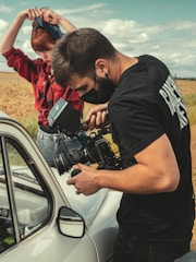 A person operates a professional video camera near a vintage car in an outdoor setting, while another person stands nearby adjusting a bandana on their head. The scene takes place in a rural environment with fields and a partly cloudy sky.