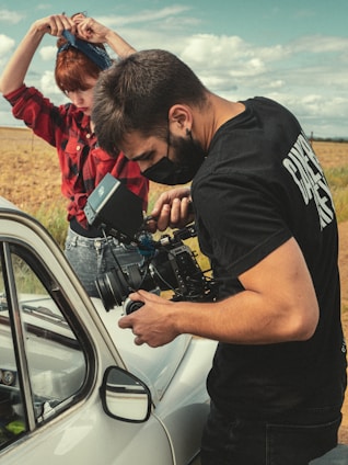 A person operates a professional video camera near a vintage car in an outdoor setting, while another person stands nearby adjusting a bandana on their head. The scene takes place in a rural environment with fields and a partly cloudy sky.