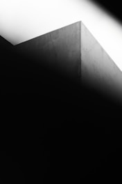 A minimalist composition featuring an angular corner of a building bathed in contrasting light and shadows. The stark geometry and interplay of light create a sense of abstraction.