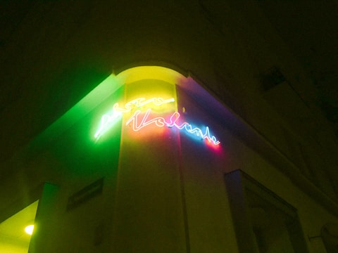 A neon sign displaying colorful, cursive text is mounted on the corner of a building wall. The sign emits a glow with a range of vibrant colors, including green, yellow, pink, and blue. This illumination casts a subtle light on the surrounding architecture during nighttime.