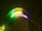 A neon sign displaying colorful, cursive text is mounted on the corner of a building wall. The sign emits a glow with a range of vibrant colors, including green, yellow, pink, and blue. This illumination casts a subtle light on the surrounding architecture during nighttime.