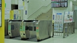 A subway station entrance features multiple turnstiles. Beyond the turnstiles, a staircase leads upward, accompanied by several signs in Japanese. An information board and timetable are visible on the right side. The environment is clean and well-lit, with a modern and organized design.