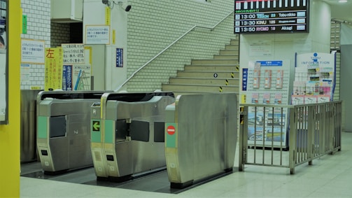 A subway station entrance features multiple turnstiles. Beyond the turnstiles, a staircase leads upward, accompanied by several signs in Japanese. An information board and timetable are visible on the right side. The environment is clean and well-lit, with a modern and organized design.