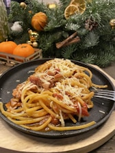 A plate of spaghetti with tomato sauce and grated cheese is set on a black plate. There is a garnish of meat pieces mixed within the pasta. In the background, a festive arrangement includes pine branches, dried orange slices, cinnamon sticks, and small orange fruits. The scene suggests a holiday or celebratory setting.