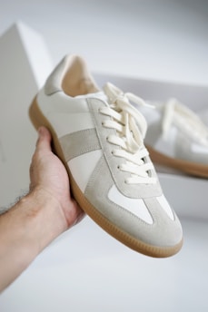 A hand holding a stylish sneaker, featuring a combination of suede and leather materials in light gray and white colors. The sneaker has clean lines, a rubber sole, and is positioned against a blurred background that includes another similar shoe in a box.