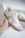 A hand holding a stylish sneaker, featuring a combination of suede and leather materials in light gray and white colors. The sneaker has clean lines, a rubber sole, and is positioned against a blurred background that includes another similar shoe in a box.