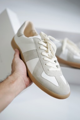 A hand holding a stylish sneaker, featuring a combination of suede and leather materials in light gray and white colors. The sneaker has clean lines, a rubber sole, and is positioned against a blurred background that includes another similar shoe in a box.