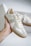 A hand holding a stylish sneaker, featuring a combination of suede and leather materials in light gray and white colors. The sneaker has clean lines, a rubber sole, and is positioned against a blurred background that includes another similar shoe in a box.