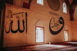 Large calligraphy adorns the walls of a spacious room with arched windows that allow sunlight to stream in. The walls feature intricate Arabic script in bold black on a neutral background. The floor is covered with a richly patterned carpet in shades of red and blue, adding to the serene ambiance.