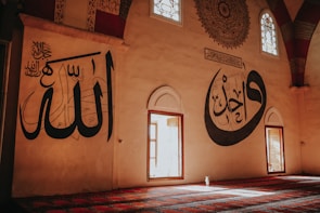 Large calligraphy adorns the walls of a spacious room with arched windows that allow sunlight to stream in. The walls feature intricate Arabic script in bold black on a neutral background. The floor is covered with a richly patterned carpet in shades of red and blue, adding to the serene ambiance.