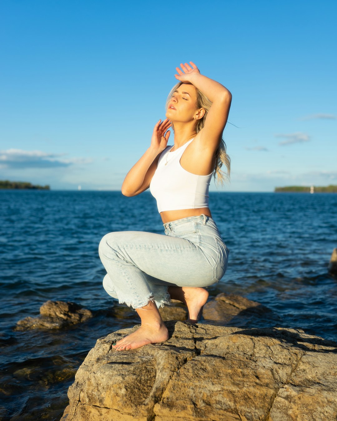 woman in white tank top and gray pants sitting on rock near body of water during