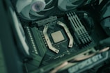 A close-up view of a computer's motherboard featuring a central processing unit (CPU) surrounded by metallic brackets. Several RAM sticks are located to the right, while cooling fans are placed above. The image captures the intricate components and layout of modern computer hardware.