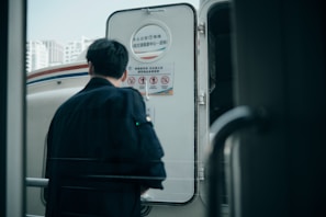 A person is seen from behind, wearing a dark coat, standing in front of the entrance to a vehicle. The door displays information and warning signs in a language that appears to be Asian. The scene is urban, with buildings visible in the background, suggesting a city setting.