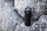 A black water bottle with a loop handle rests against rough gray rocks. The bottle has a clear cap, indicating it may be used for outdoor activities or hiking.