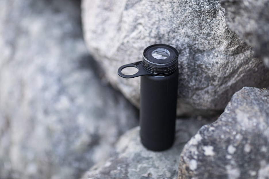 A black water bottle with a loop handle rests against rough gray rocks. The bottle has a clear cap, indicating it may be used for outdoor activities or hiking.