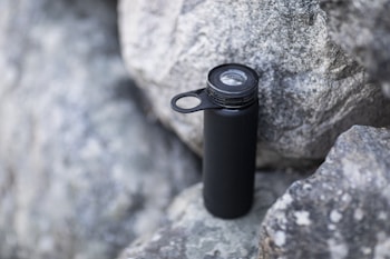 A black water bottle with a loop handle rests against rough gray rocks. The bottle has a clear cap, indicating it may be used for outdoor activities or hiking.