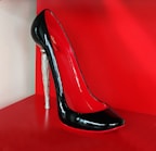 A high-heeled shoe with a glossy black exterior and red interior, featuring a unique silver stiletto heel. The shoe is displayed against a bright red background, highlighting its elegant and sophisticated design.