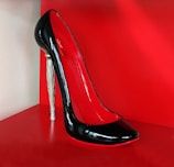 A high-heeled shoe with a glossy black exterior and red interior, featuring a unique silver stiletto heel. The shoe is displayed against a bright red background, highlighting its elegant and sophisticated design.