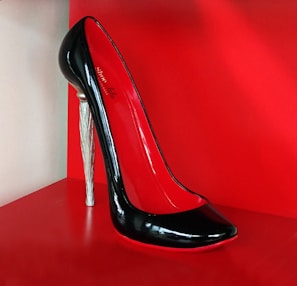 A high-heeled shoe with a glossy black exterior and red interior, featuring a unique silver stiletto heel. The shoe is displayed against a bright red background, highlighting its elegant and sophisticated design.