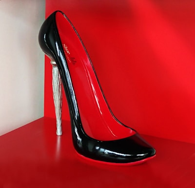 A high-heeled shoe with a glossy black exterior and red interior, featuring a unique silver stiletto heel. The shoe is displayed against a bright red background, highlighting its elegant and sophisticated design.